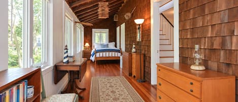 A bedroom with a wooden ceiling, a bed with a blue and white bedspread, a nightstand with a lamp, a dresser, and a bookshelf with books.