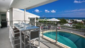 Un espace aménagé au bord de la piscine avec une table et des chaises, une piscine fermée par des parois en verre, et une vue sur l’océan et les bâtiments situés au loin.