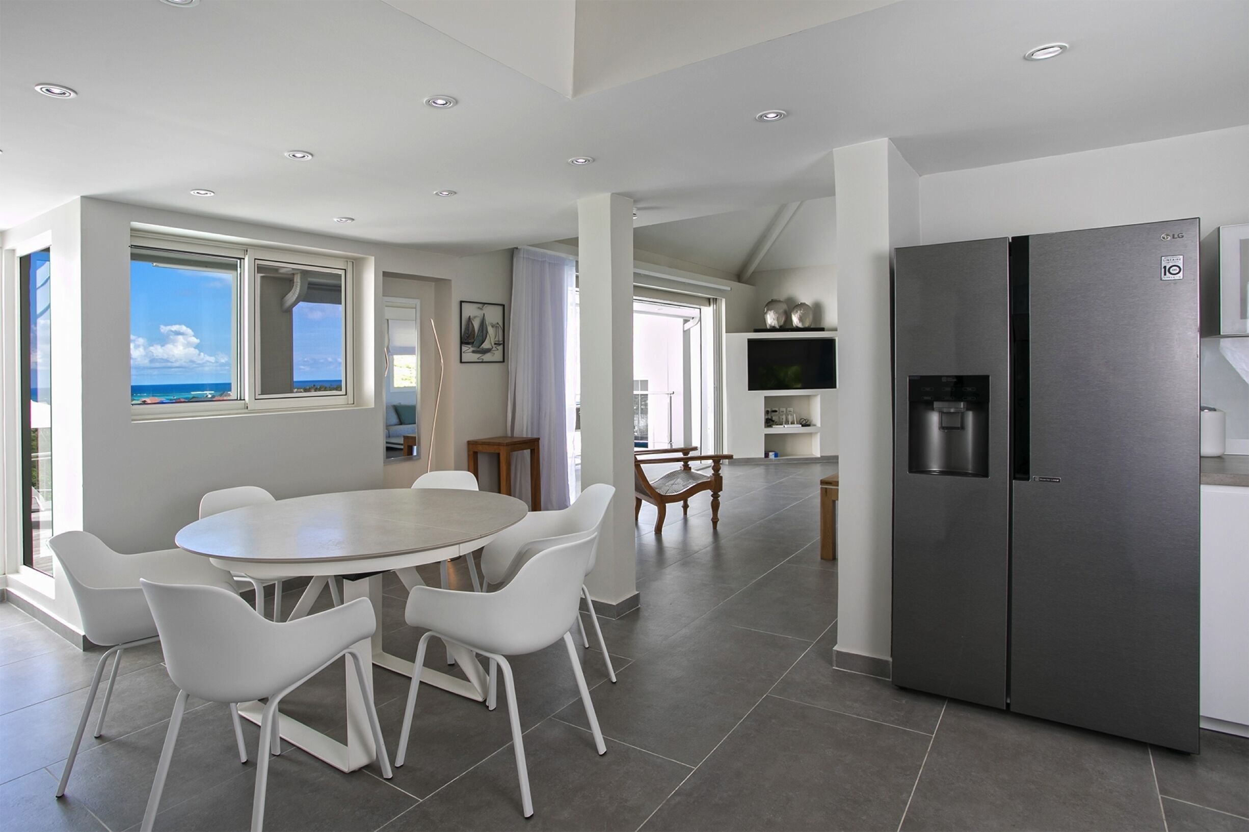A modern kitchen with a large refrigerator, white chairs around a dining table, and a view of the ocean through large windows.