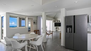 Une cuisine moderne dotée d’un grand réfrigérateur, de chaises blanches disposées autour d’une table à manger, et offrant une vue sur l’océan grâce à de grandes fenêtres.