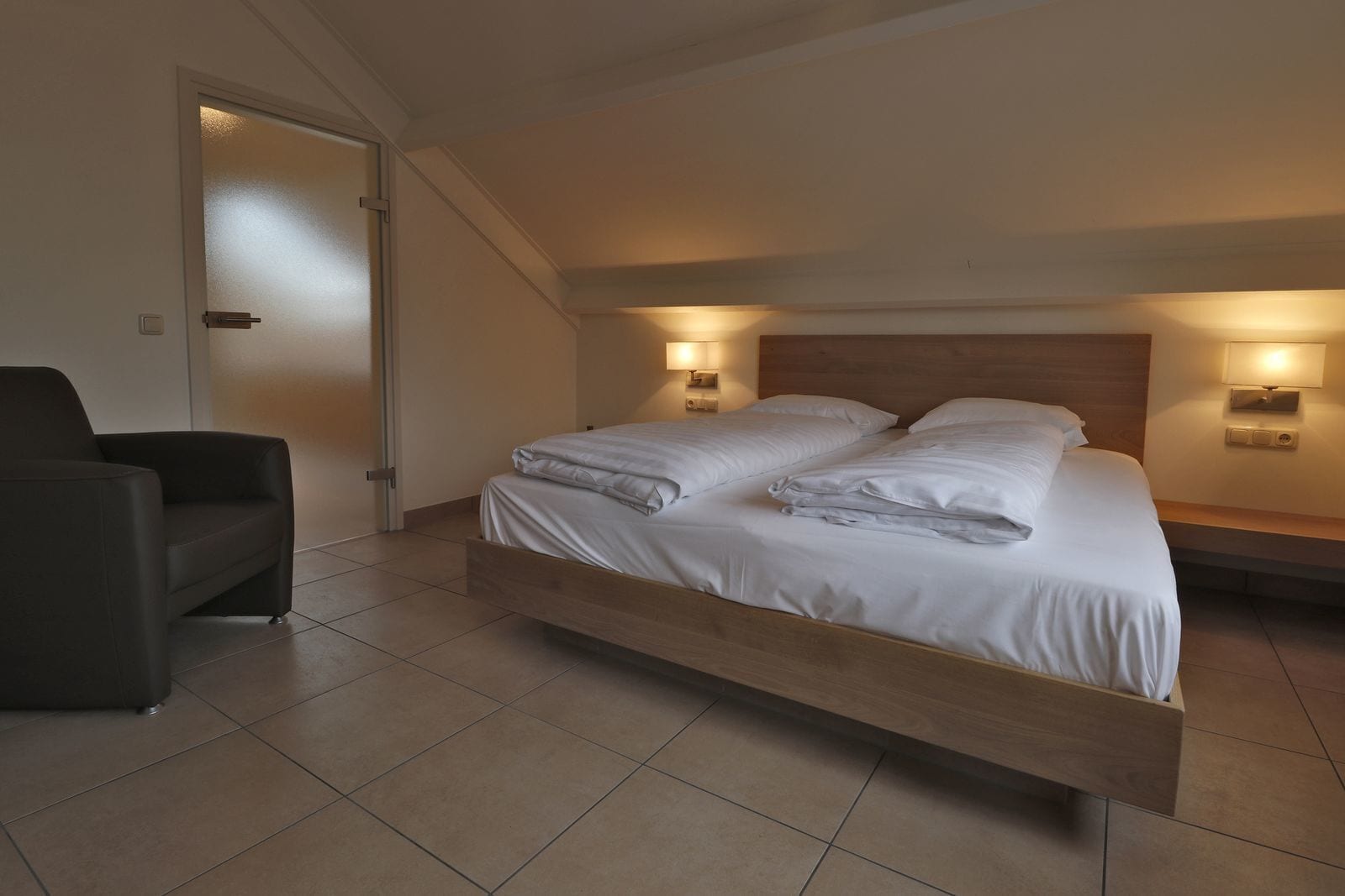 Un dormitorio moderno con una cama de madera, dos lámparas de noche, un sillón oscuro y una puerta que da a otra habitación.