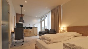 Habitación de hotel con cama, escritorio, silla, kitchenette y ventana con cortinas.