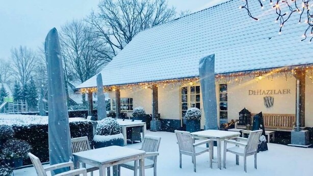 Zona de estar al aire libre cubierta de nieve con mesas y sillas, un edificio con la señalización 'DEHAZELAE' y plantas cubiertas de nieve.