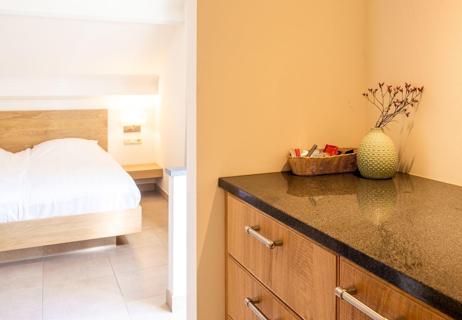 A hotel room with a bed, a nightstand, a lamp, a basket with toiletries, and a vase with dried flowers on a counter.