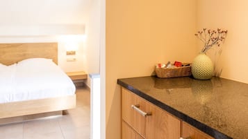 A hotel room with a bed, a nightstand, a lamp, a basket with toiletries, and a vase with dried flowers on a counter.