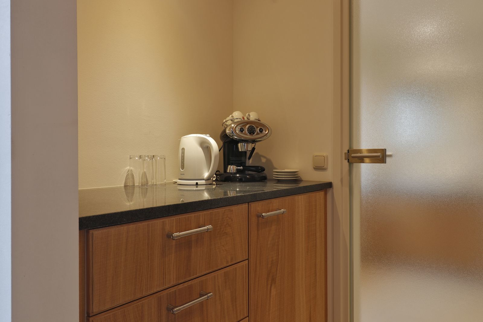 Una kitchenette con una cafetera, un hervidor y una pila de platos sobre la encimera.