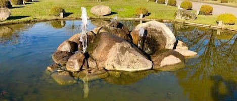 A pond with rocks and a small waterfall, surrounded by neatly trimmed bushes and trees.