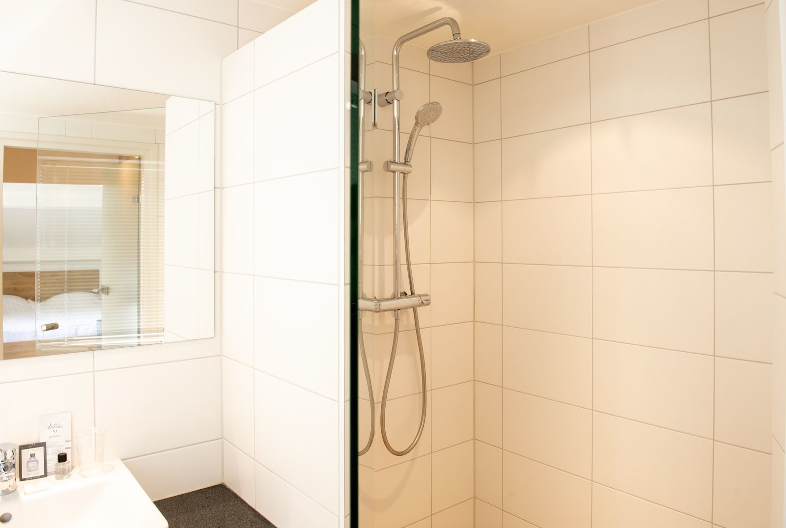 A modern bathroom with white tiles, a glass shower enclosure, and a visible bed in the adjacent room.