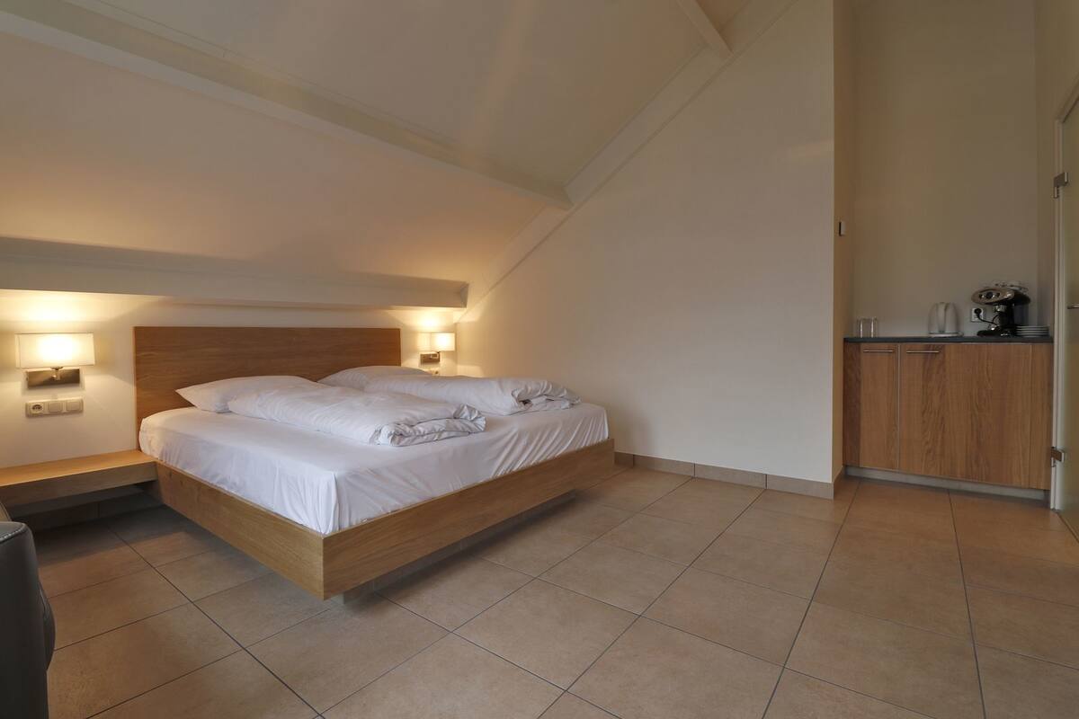 A bedroom with a wooden bed, two bedside lamps, a small bedside table, and a kitchenette area with a coffee maker.