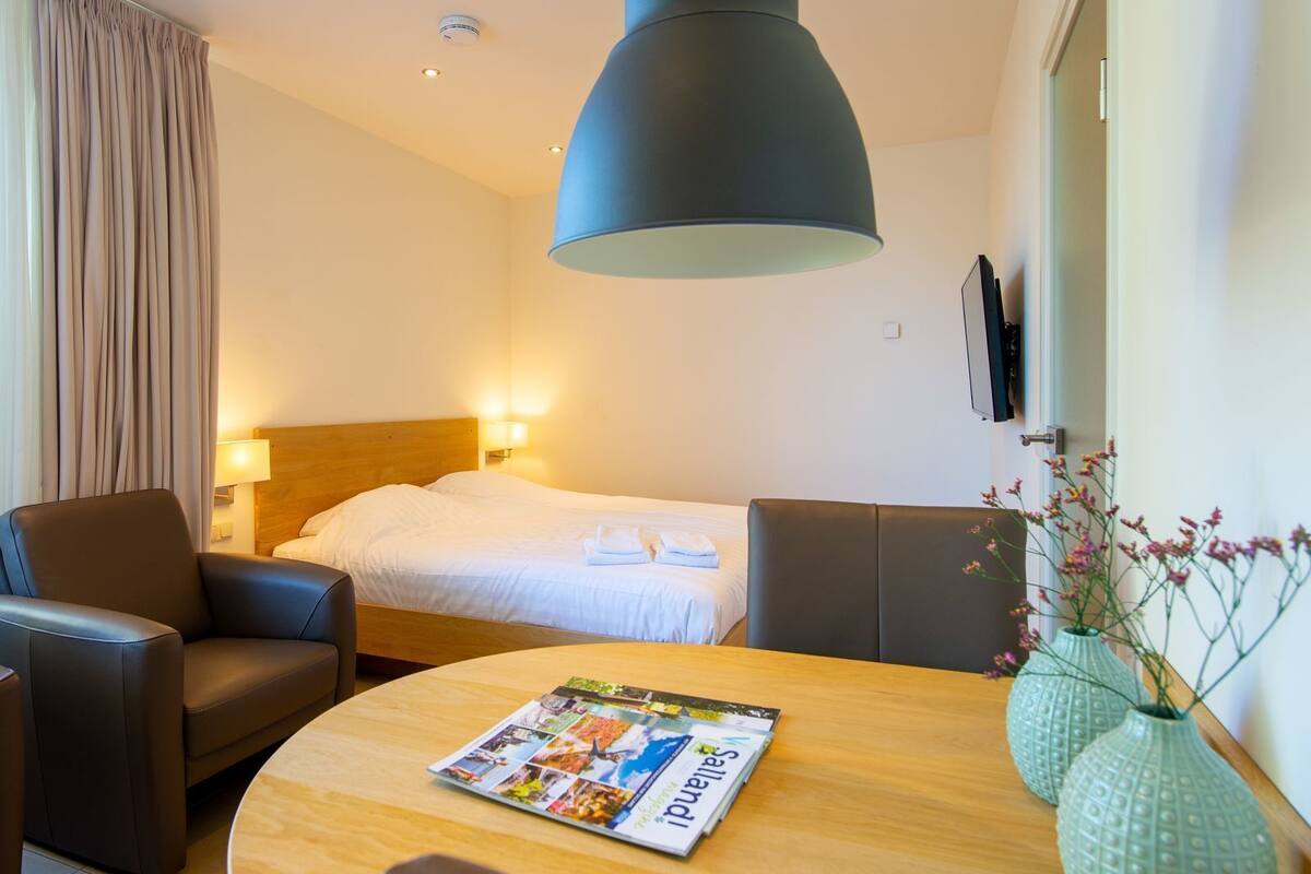 A hotel room with a bed, a desk, a chair, a lamp, and a vase with flowers.