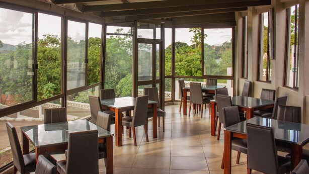 A spacious dining area with large windows offering a view of lush greenery. Tables and chairs are arranged for dining. - Cedar Place (Blantyre)