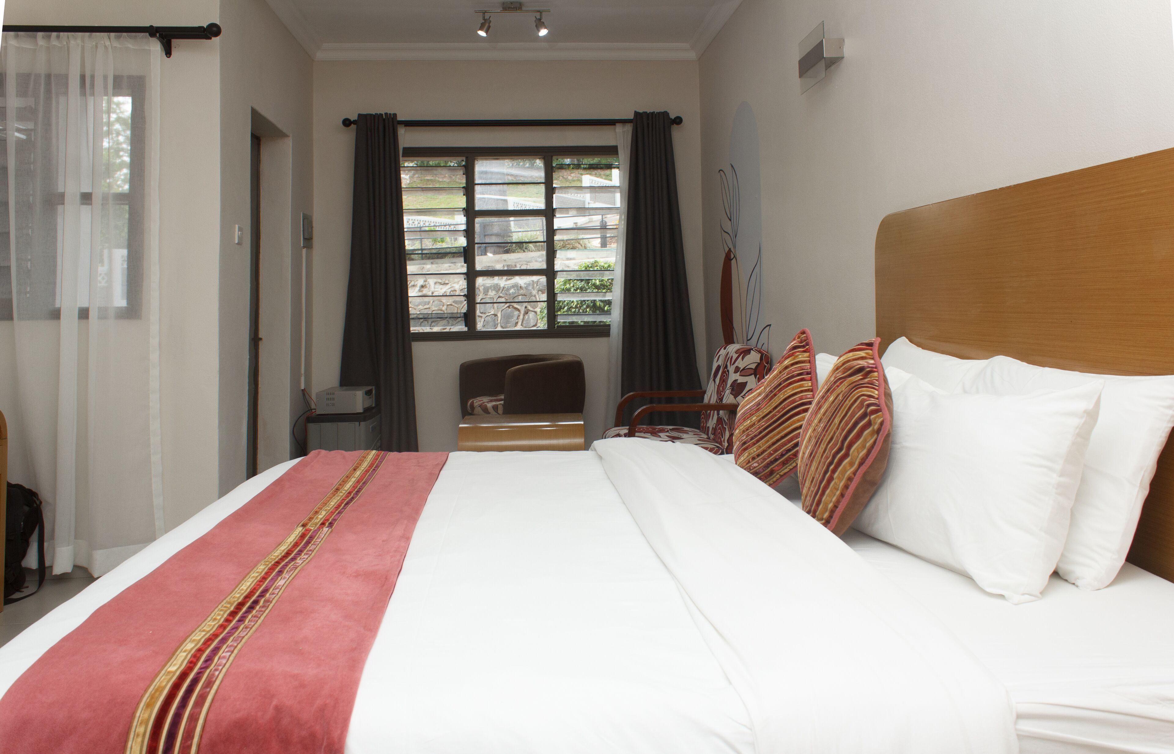 A hotel room with a large bed, a wooden headboard, and a view of the outdoors through a window with curtains.