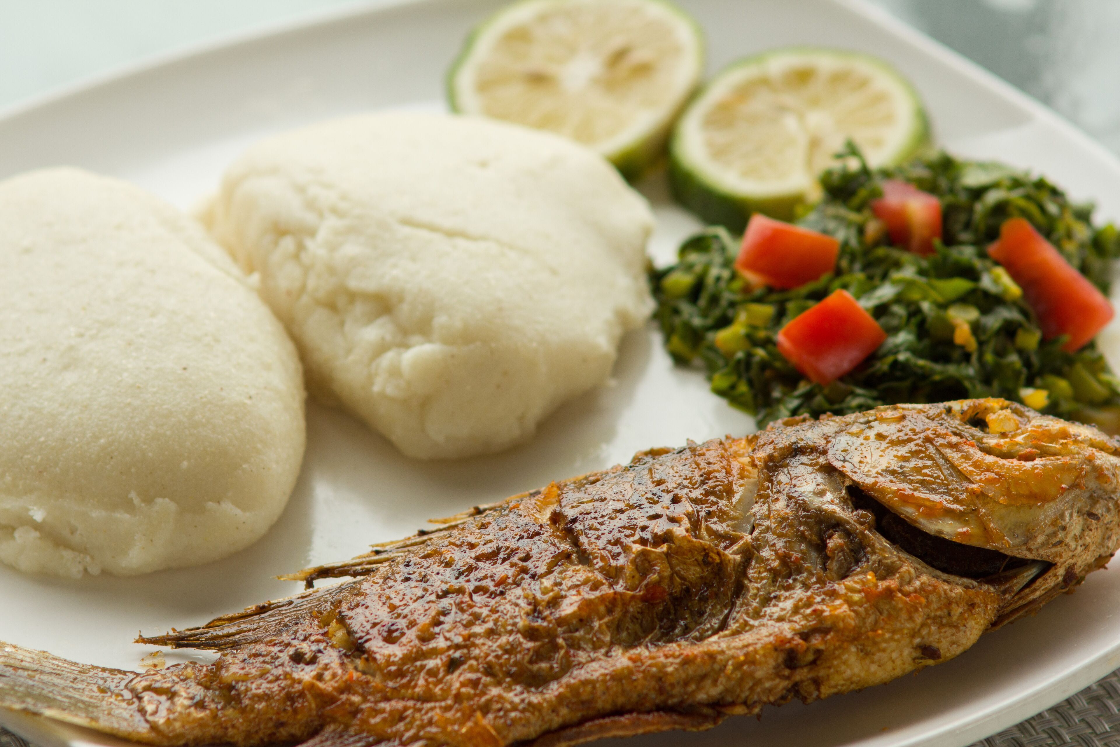 A plate with a whole fried fish, two pieces of white bread, and a side of greens and tomatoes.