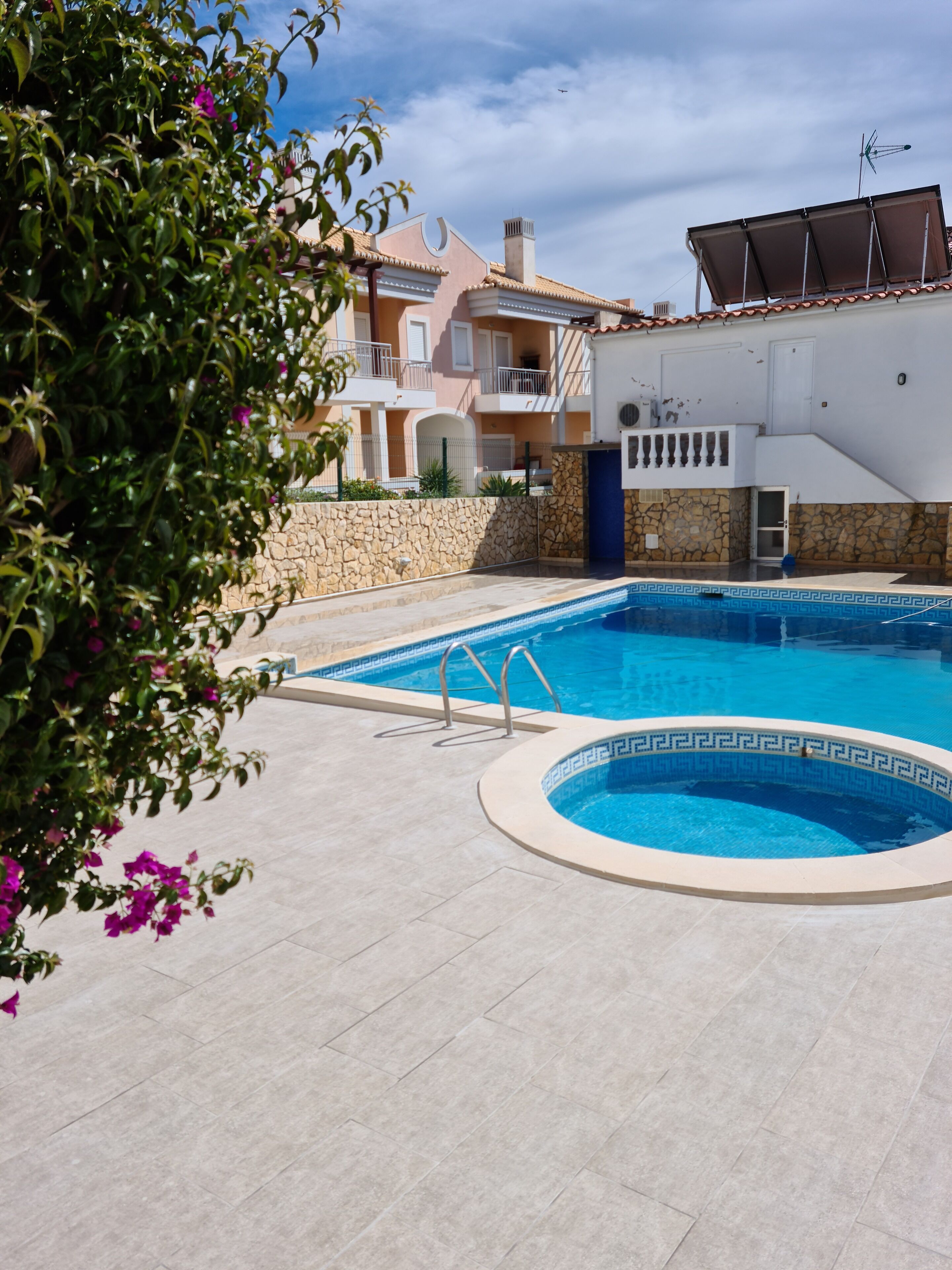 A swimming pool with a stone border, a stone wall, and a building with balconies in the background.