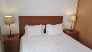 Een hotelkamer met een houten hoofdeinde, twee nachtkastjes, een bed met witte lakens en twee lampen op de tafels.