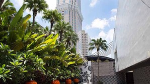 Un sendero bordeado por plantas en macetas y palmeras, que conduce a un edificio moderno con un rascacielos al fondo.
