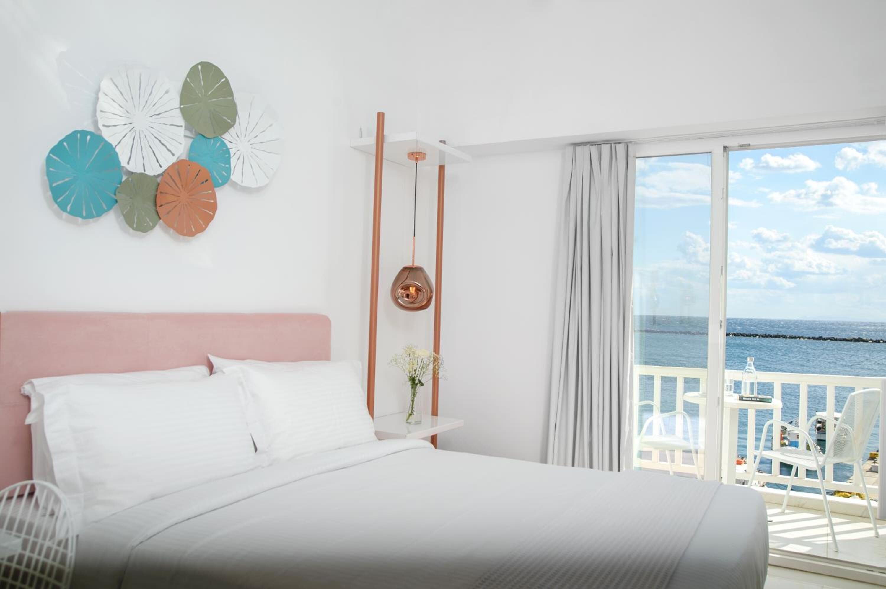Ein Hotelzimmer mit Bett, Nachttisch, Blumen in einer Vase, Blick aufs Meer und Balkon mit Sitzgelegenheiten.
