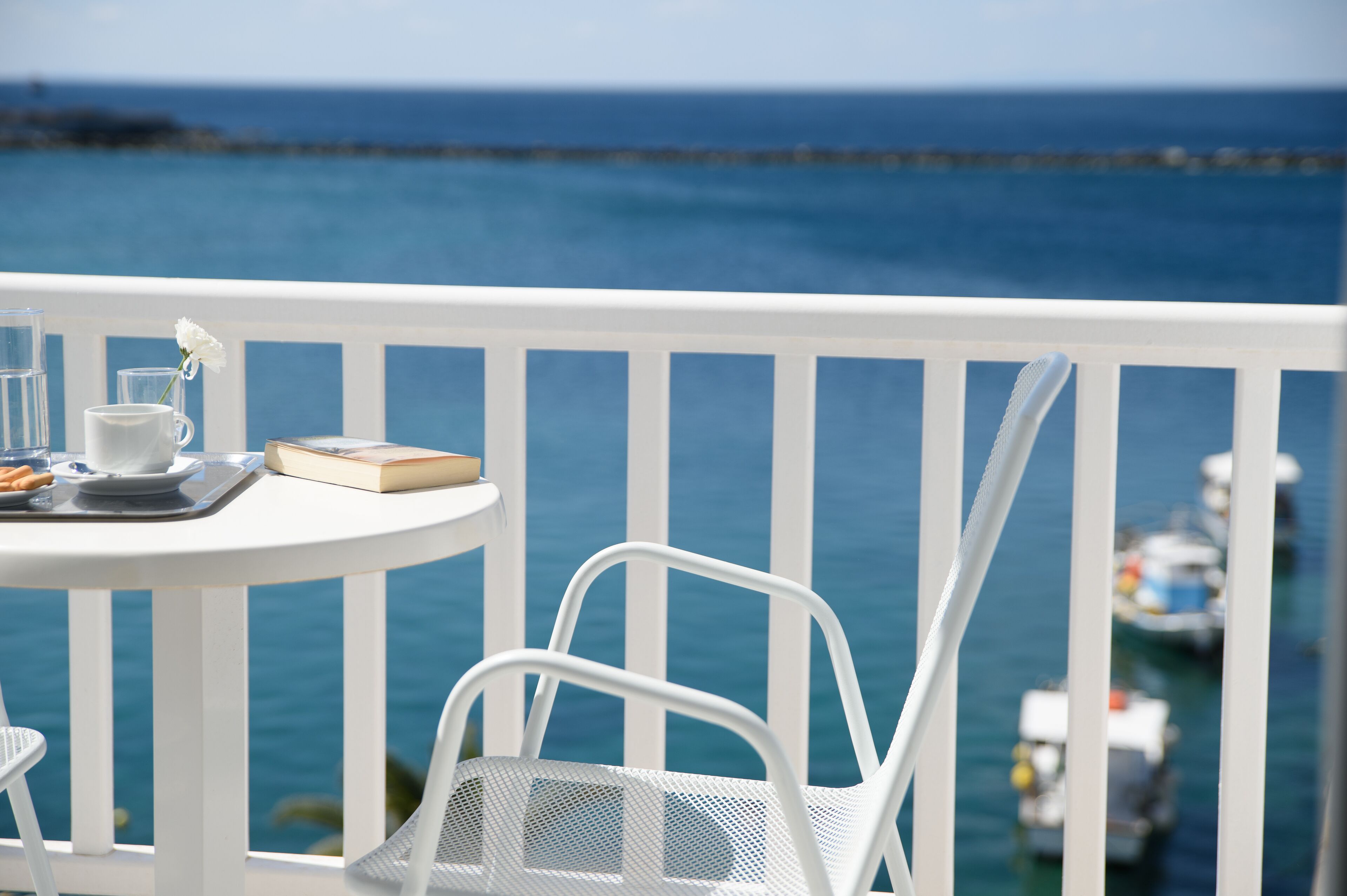 Une table en métal blanc, sur laquelle se trouvent un plateau, une tasse et un livre, est placée sur un balcon donnant sur la mer.