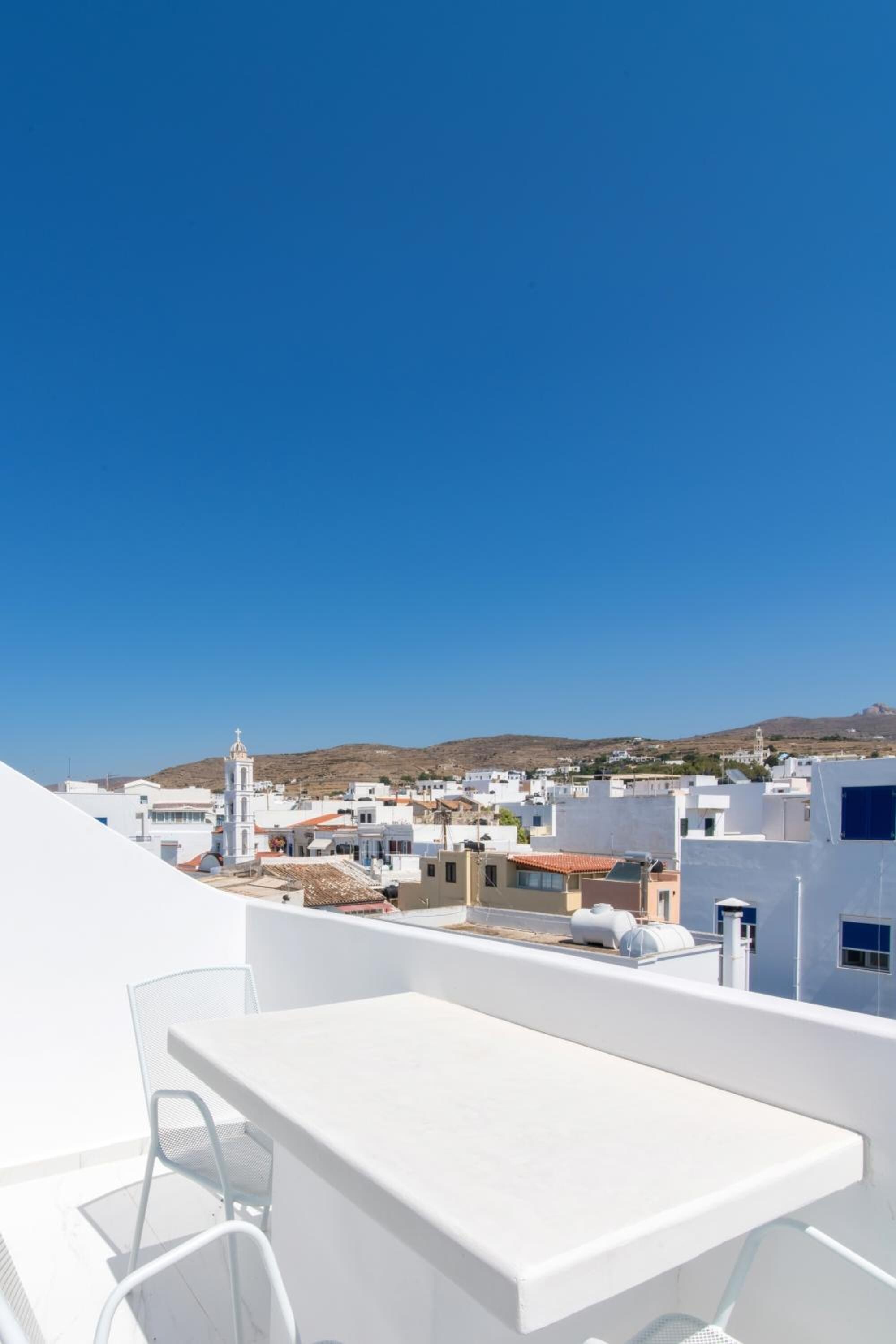 Une terrasse sur le toit, avec une table et des chaises blanches, d’où l’on peut admirer une ville côtière aux bâtiments blancs, sous un ciel bleu et dégagé.