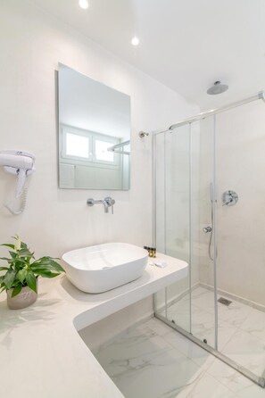 A modern bathroom with a white sink, a glass shower enclosure, and a hairdryer mounted on the wall. - Nama Boutique Hotel (Tinos)