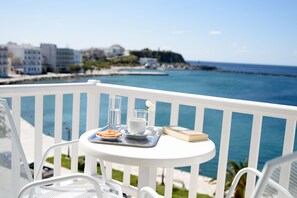 A balcony with a white railing, a small round table with a tray holding a glass of water, a cup, a book, and a plate with food. The background features a coastal view with buildings and the sea. - Nama Boutique Hotel (Tinos)