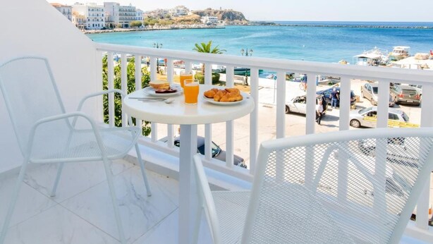 A balcony with white chairs and a table, overlooking a beach with a clear view of the sea and buildings in the distance. - Nama Boutique Hotel (Tinos)