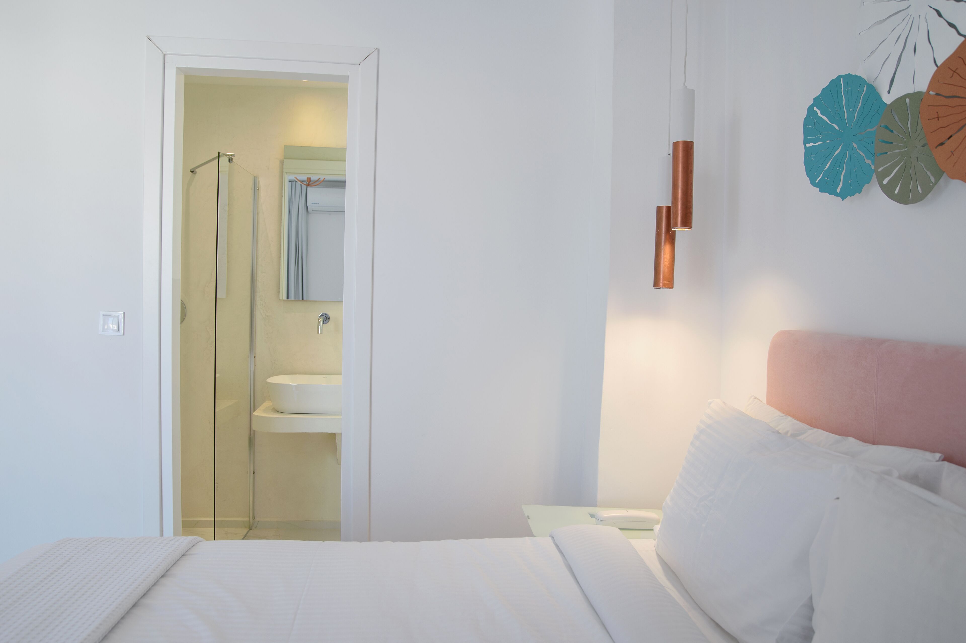 A bedroom with a bed, a nightstand, a bedside table, a wall-mounted lamp, and a bathroom visible through an open door.