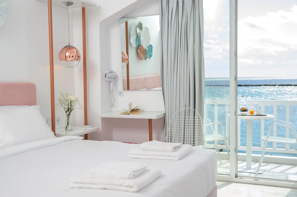 A modern hotel room with a large bed, a bedside table with a book and a vase of flowers, a mirror, and a view of the sea through a balcony. - Nama Boutique Hotel (Tinos)