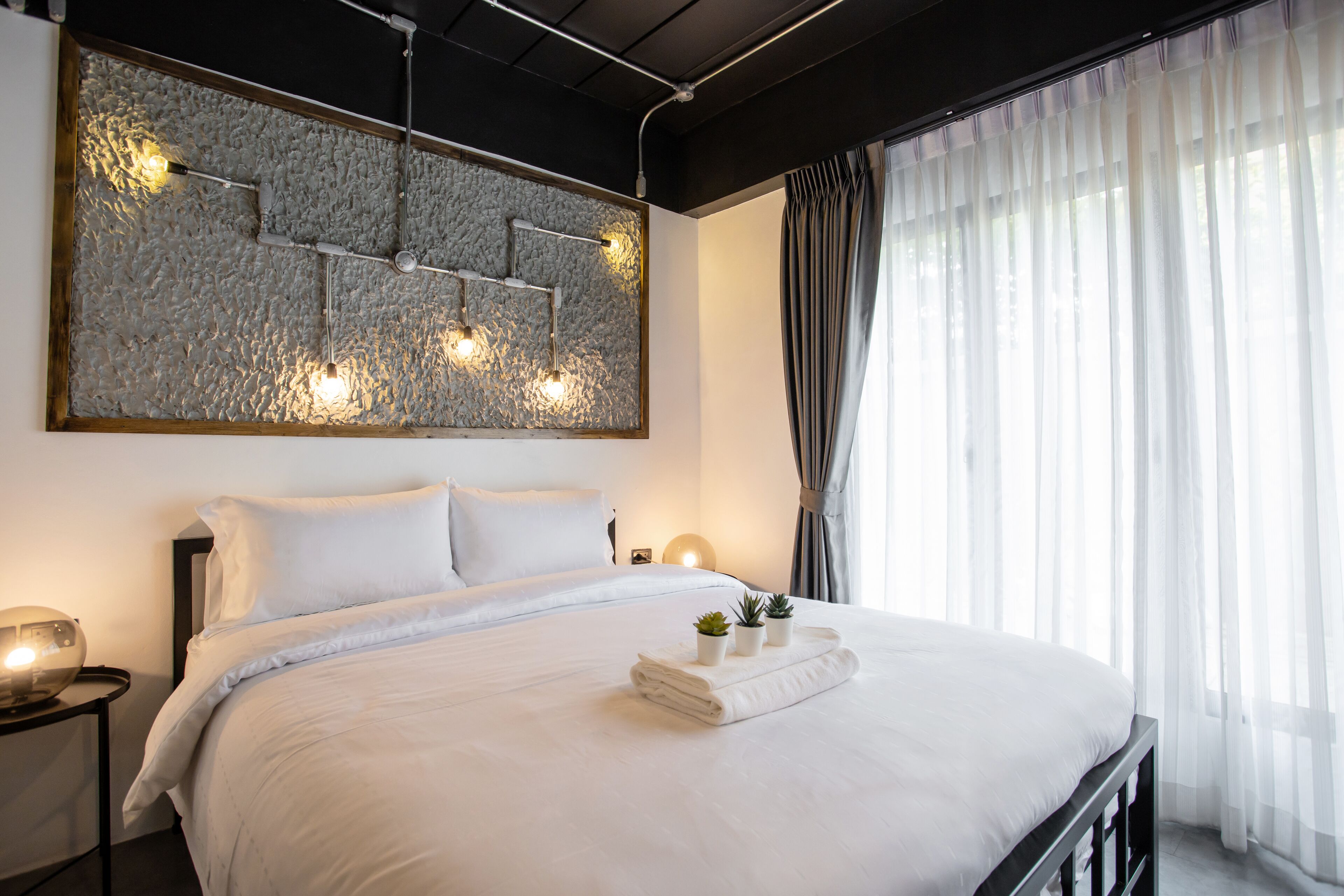 Una habitación de hotel moderna con una cama grande, un cuadro de pared con textura y un amplio ventanal con cortinas traslúcidas.