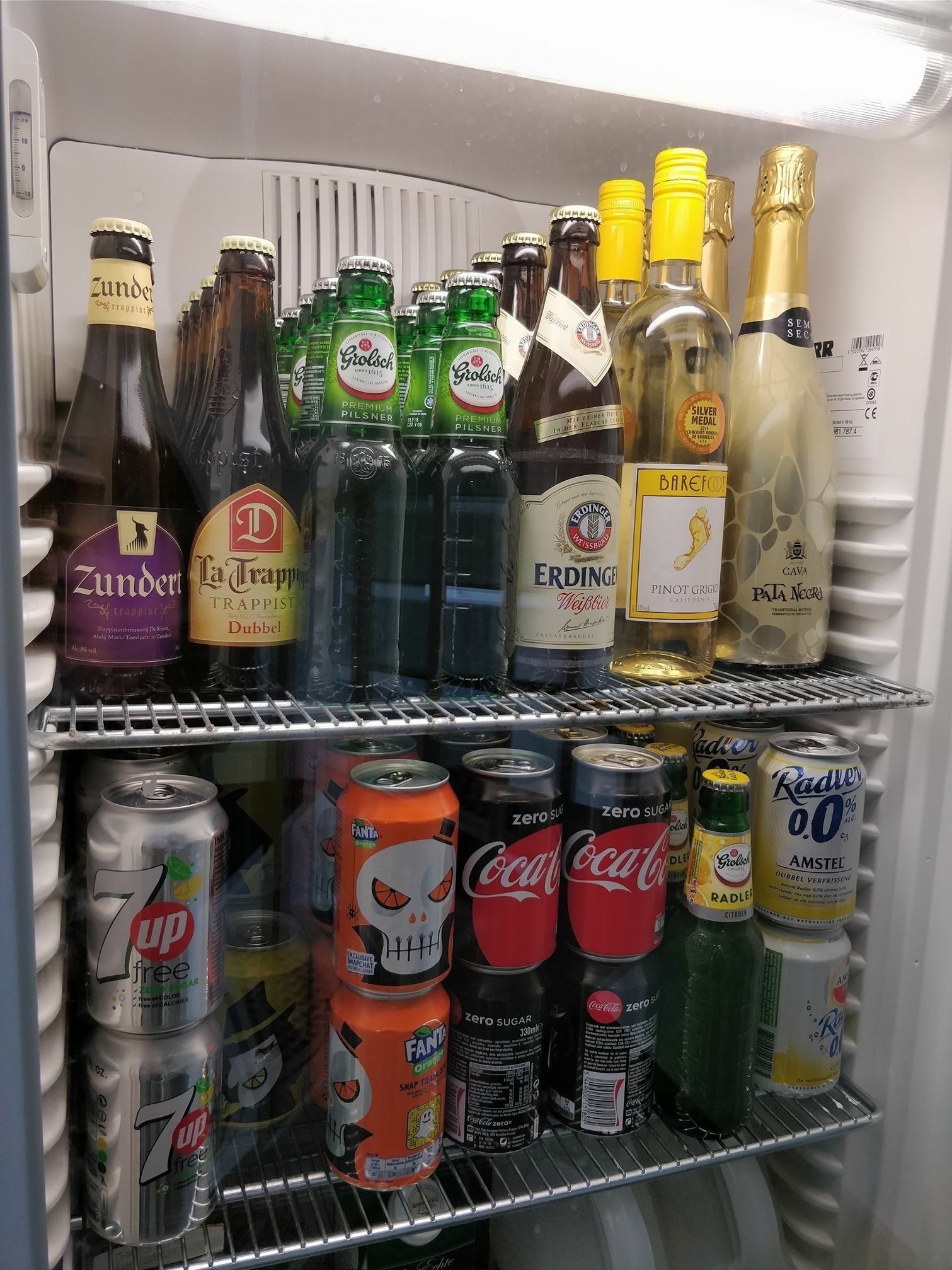 Un refrigerador lleno de diversas bebidas, incluyendo botellas de Zunder, La Trappist y Erdinger, así como latas de Fanta y Coca-Cola Zero Azúcar.