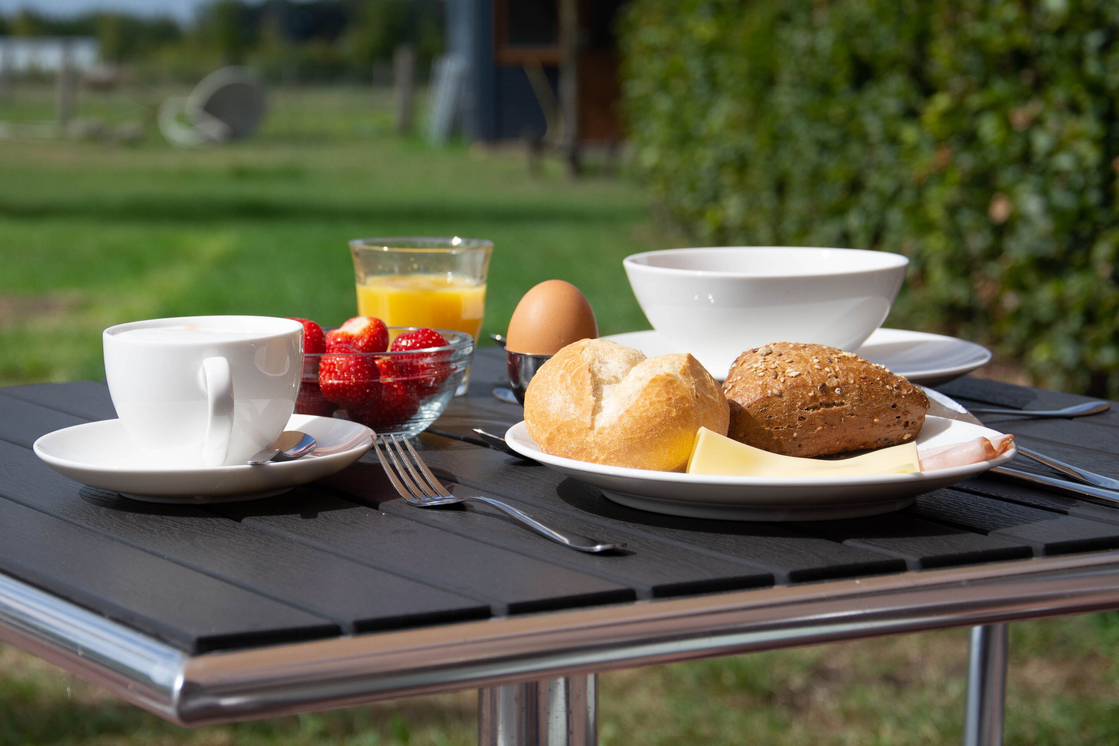 Een ontbijttafel met een kopje, kom, bord met brood, aardbeien en een glas jus d'orange.