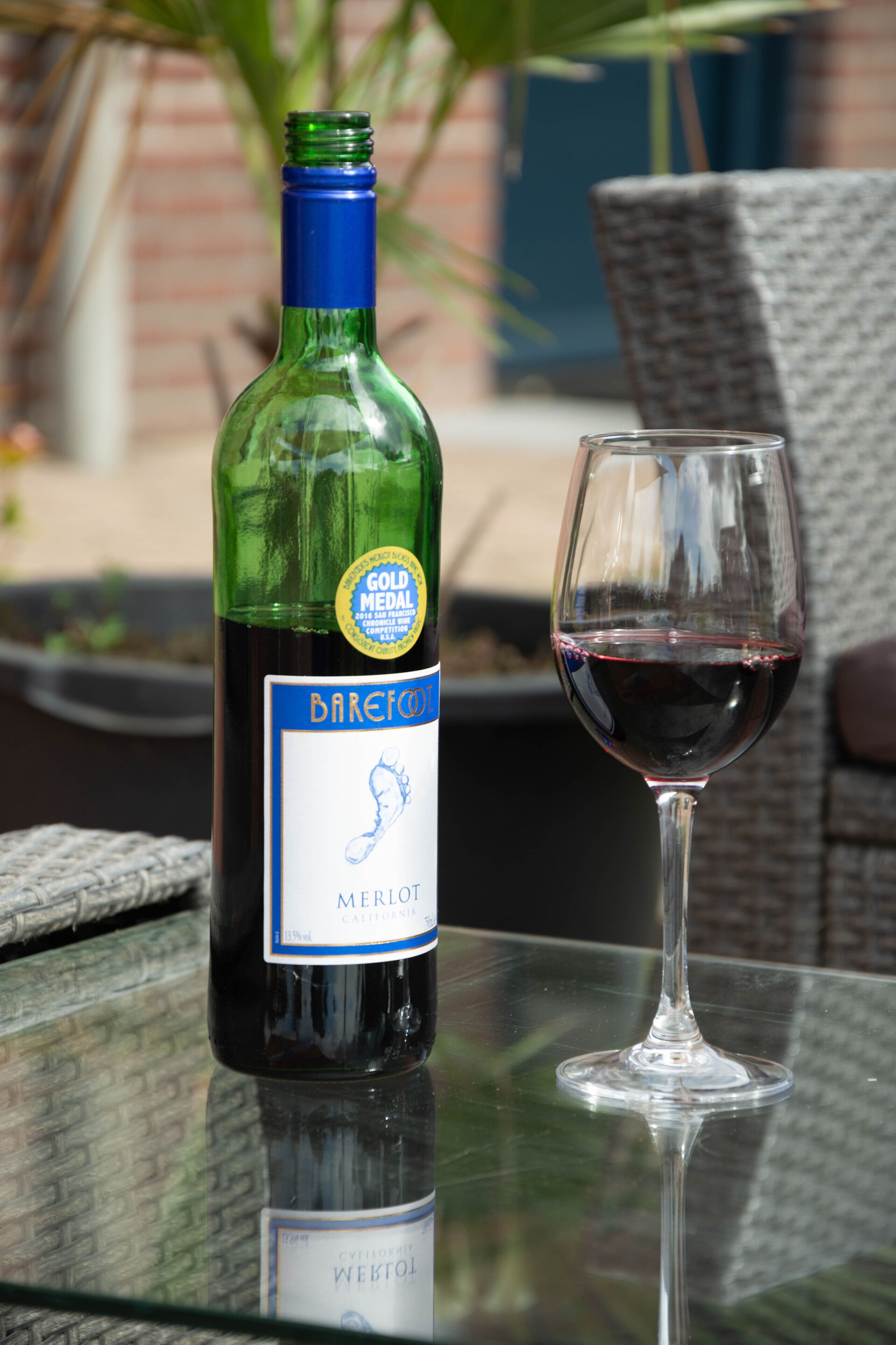 Een fles Barefoot Merlot en een wijnglas op een glazen tafel.
