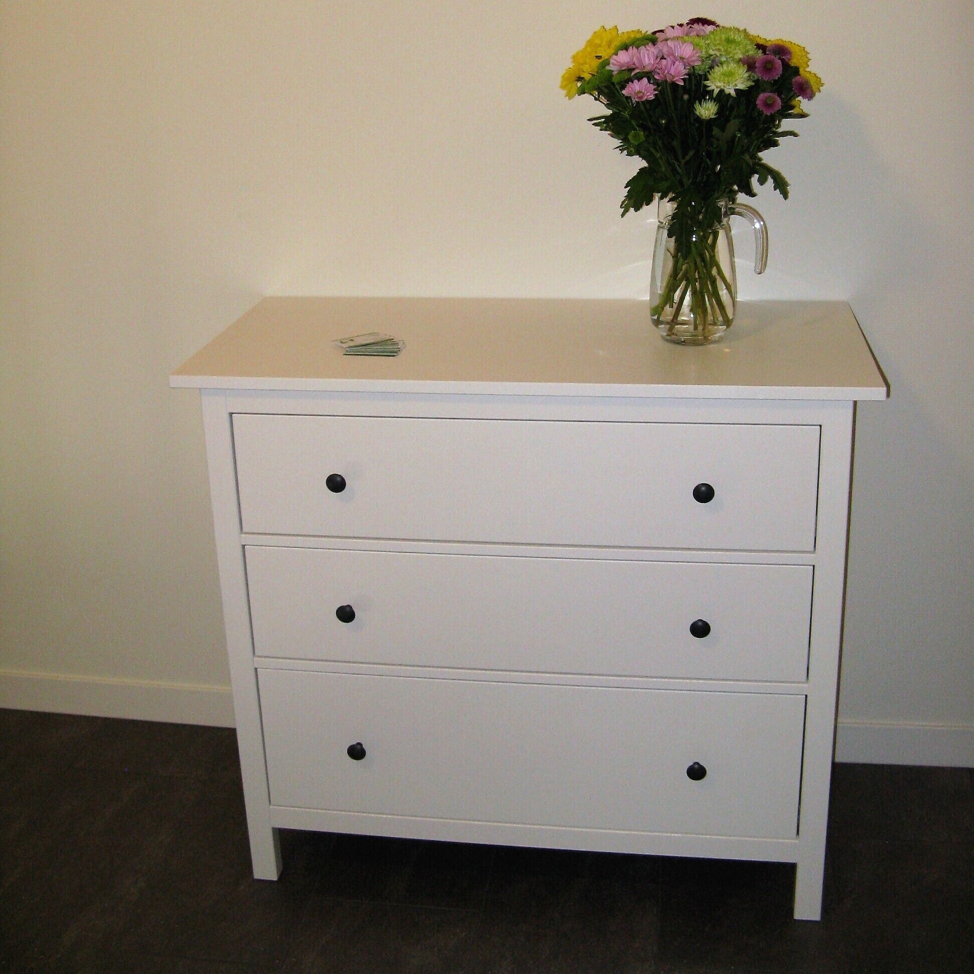 A white bedside table with three drawers and a vase of flowers on top.