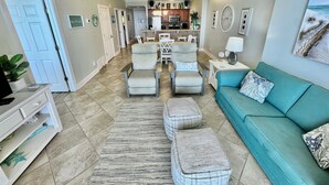 Un salon spacieux avec un canapé de couleur turquoise, deux fauteuils, une table basse et un tapis. Au fond, on aperçoit un coin cuisine avec une table à manger et des chaises.