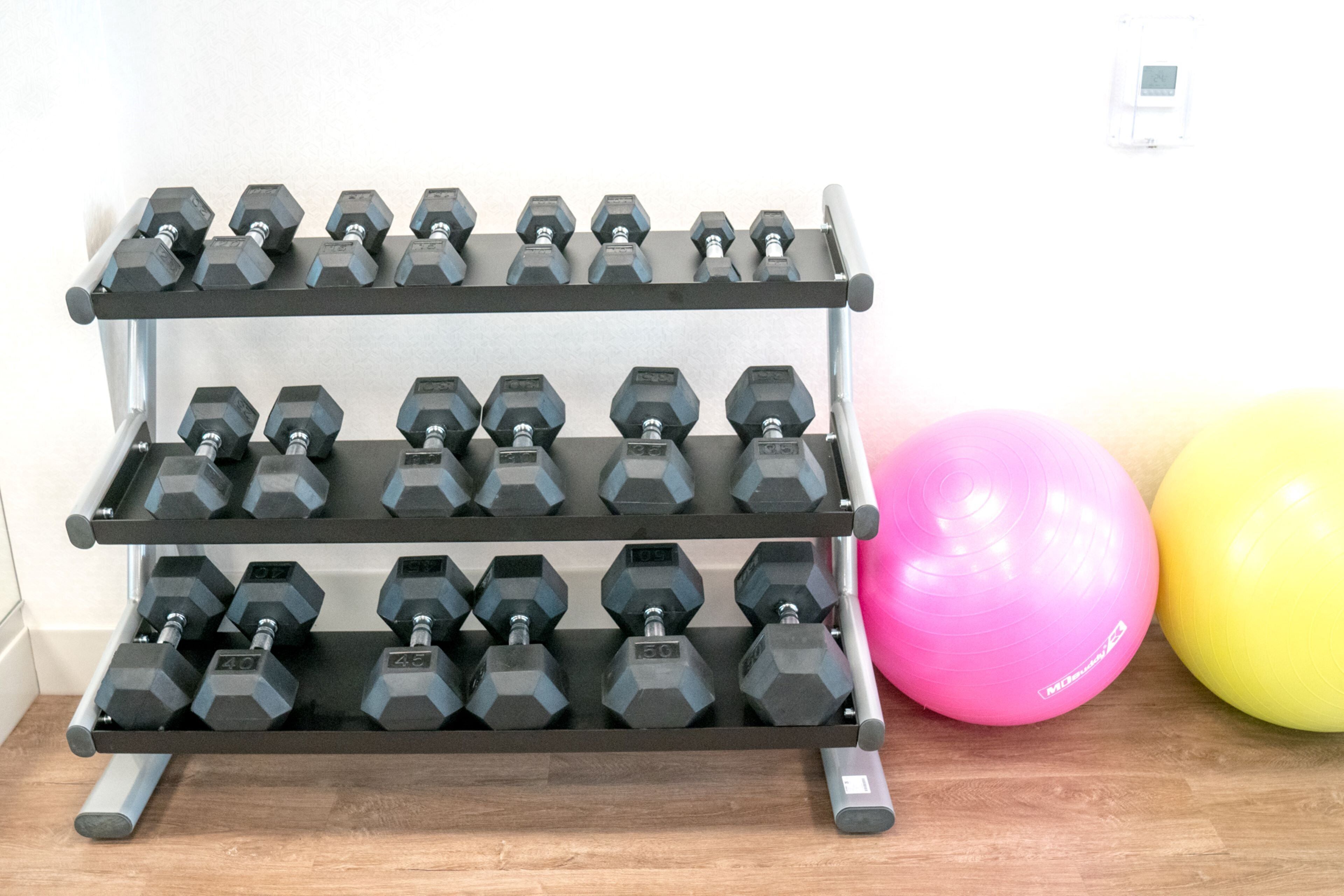 Un soporte para mancuernas de tres niveles con pesas, una pelota de ejercicios rosa y una amarilla, y una pared blanca al fondo.