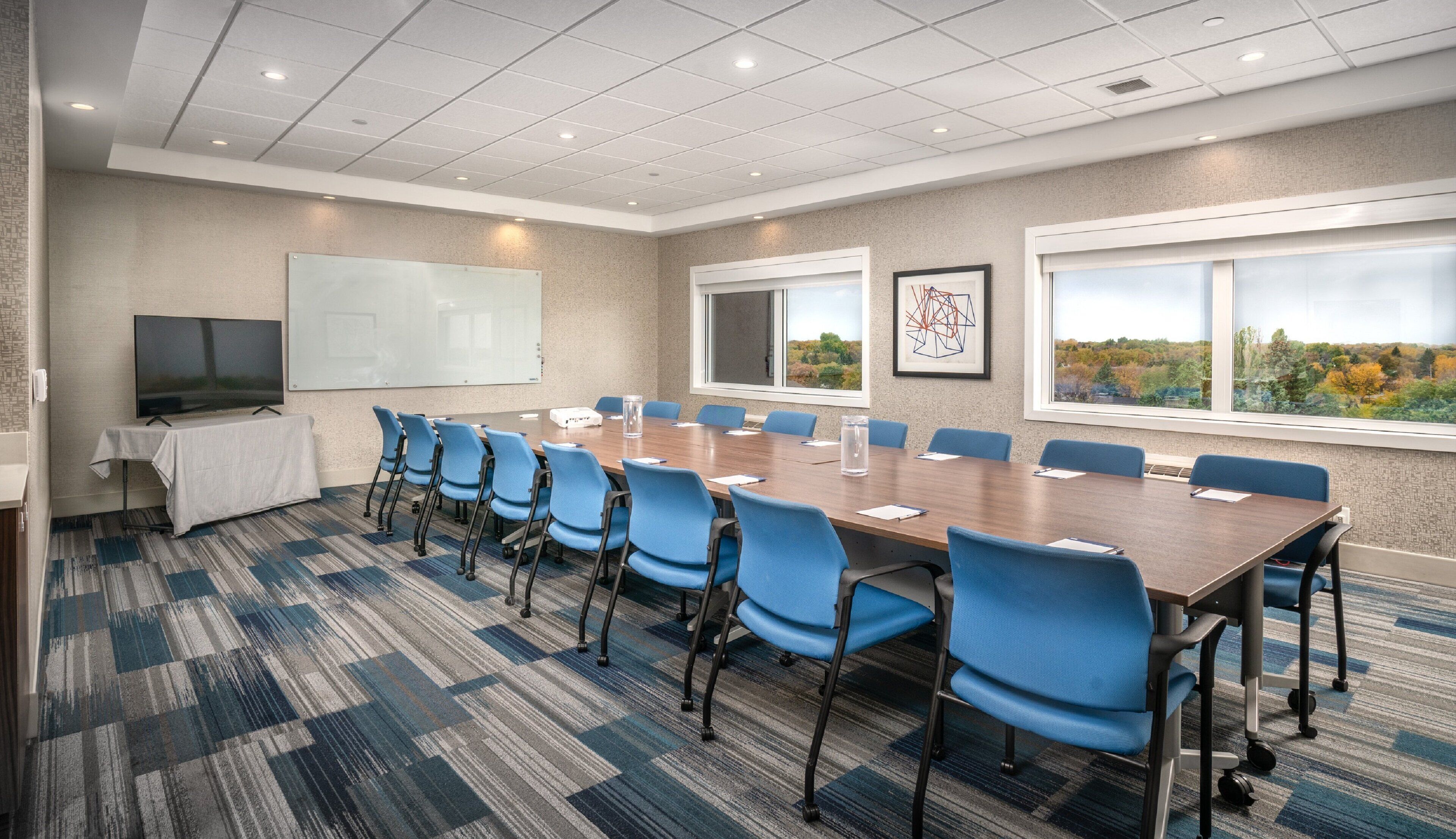 A conference room with a long table, blue chairs, a whiteboard, and a TV.