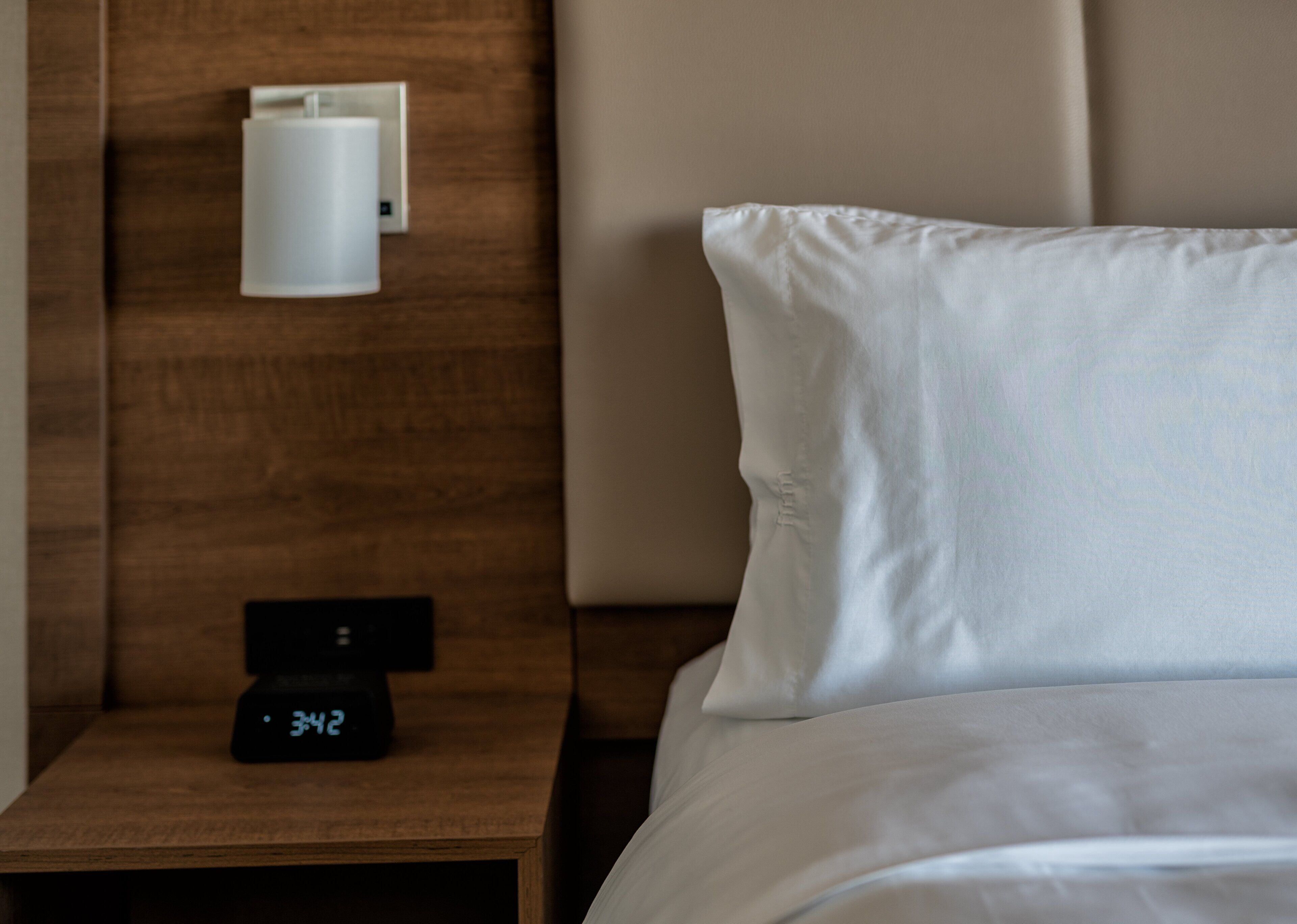 Ein ordentlich bezogenes Bett mit weißen Leinen, ein Nachttisch mit digitaler Uhr und eine wandmontierte Leuchte.