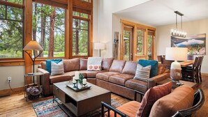 A spacious living room with a large sectional sofa, a coffee table, and a dining area with a wooden table and chairs. Large windows provide a view of the surrounding forest.