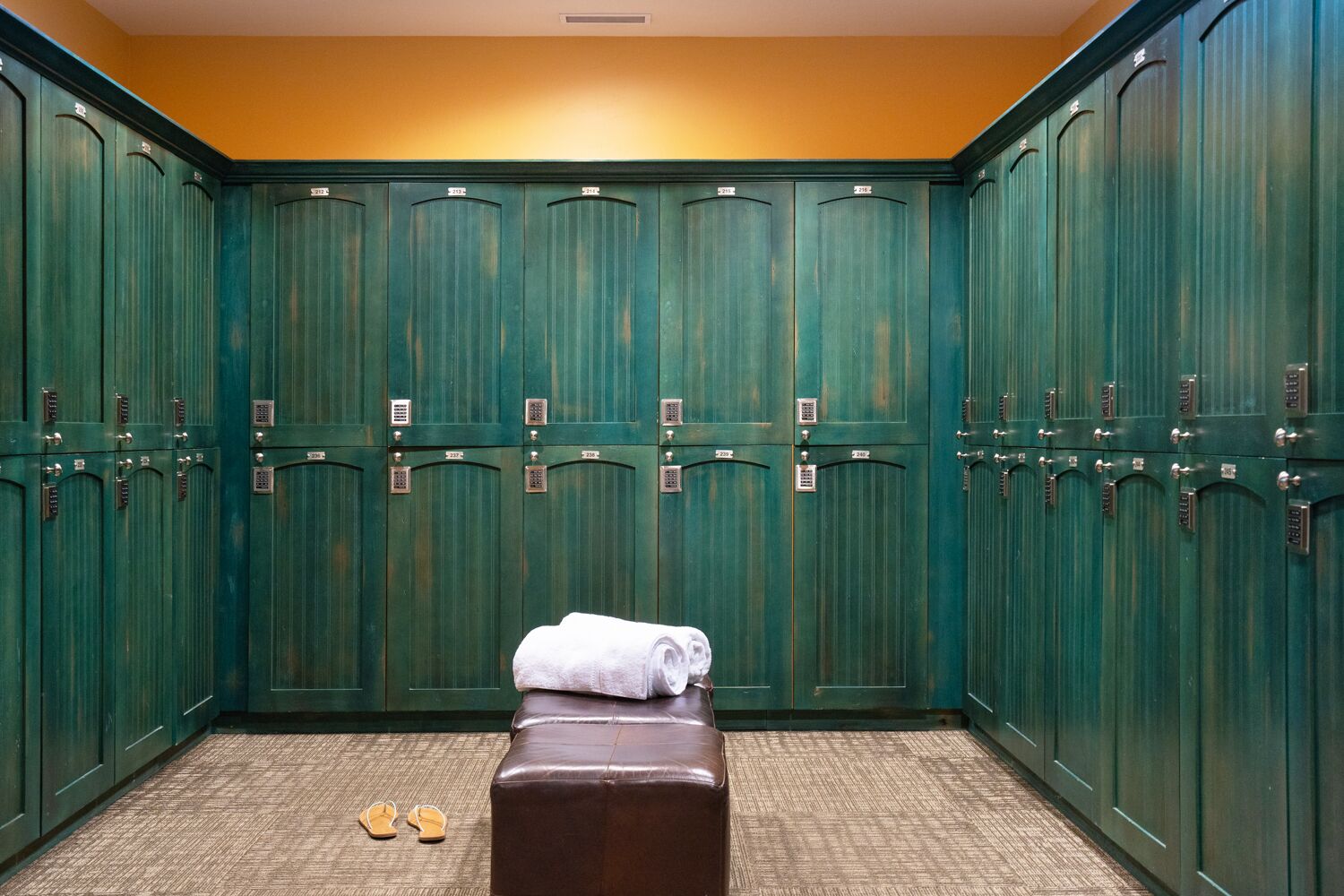 A row of green lockers with numbers on the doors, a bench with a towel, and a pair of sandals on the floor.
