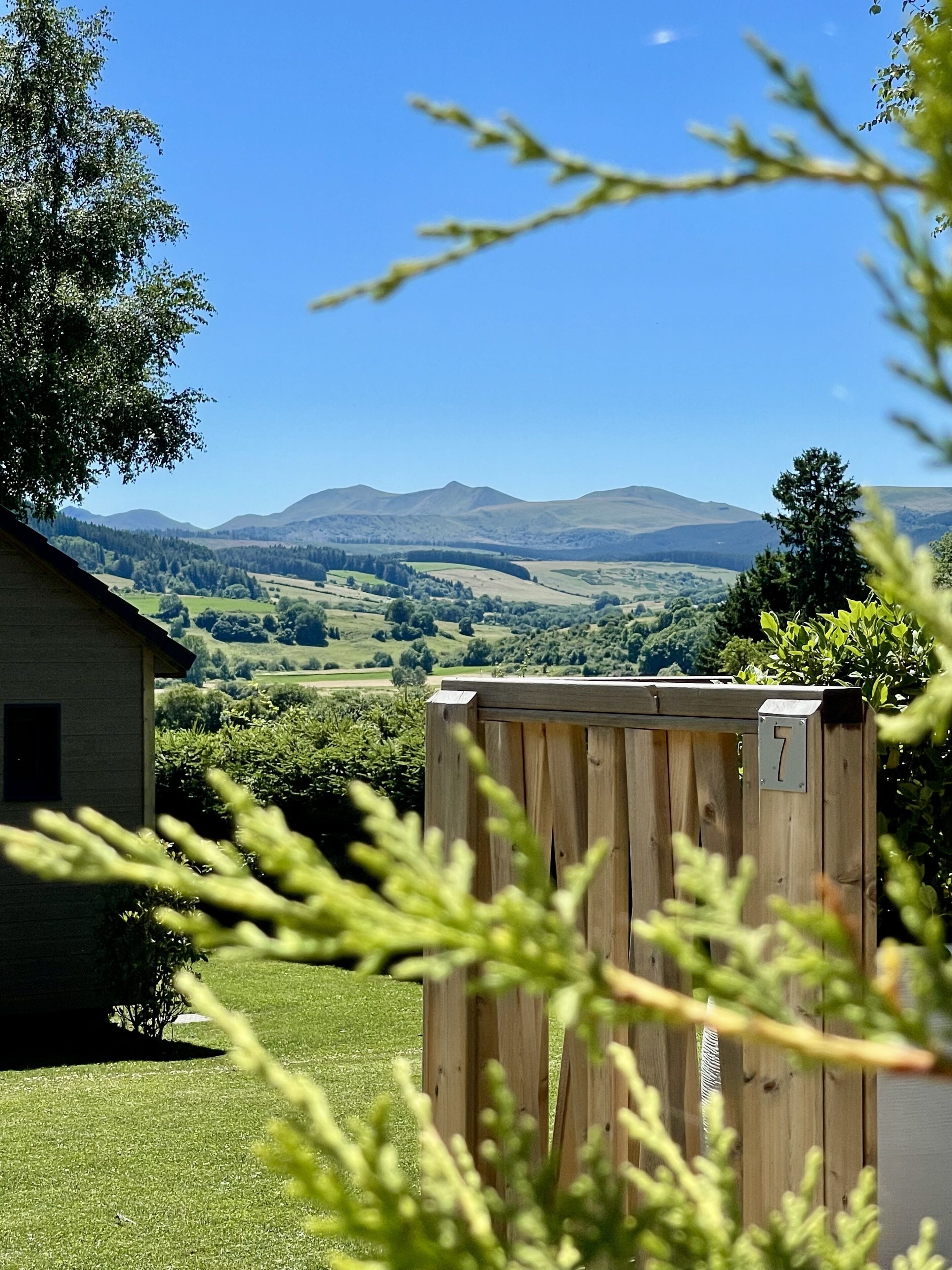 A wooden fence with a number plate in a rural setting, a house, and a view of rolling hills and mountains.