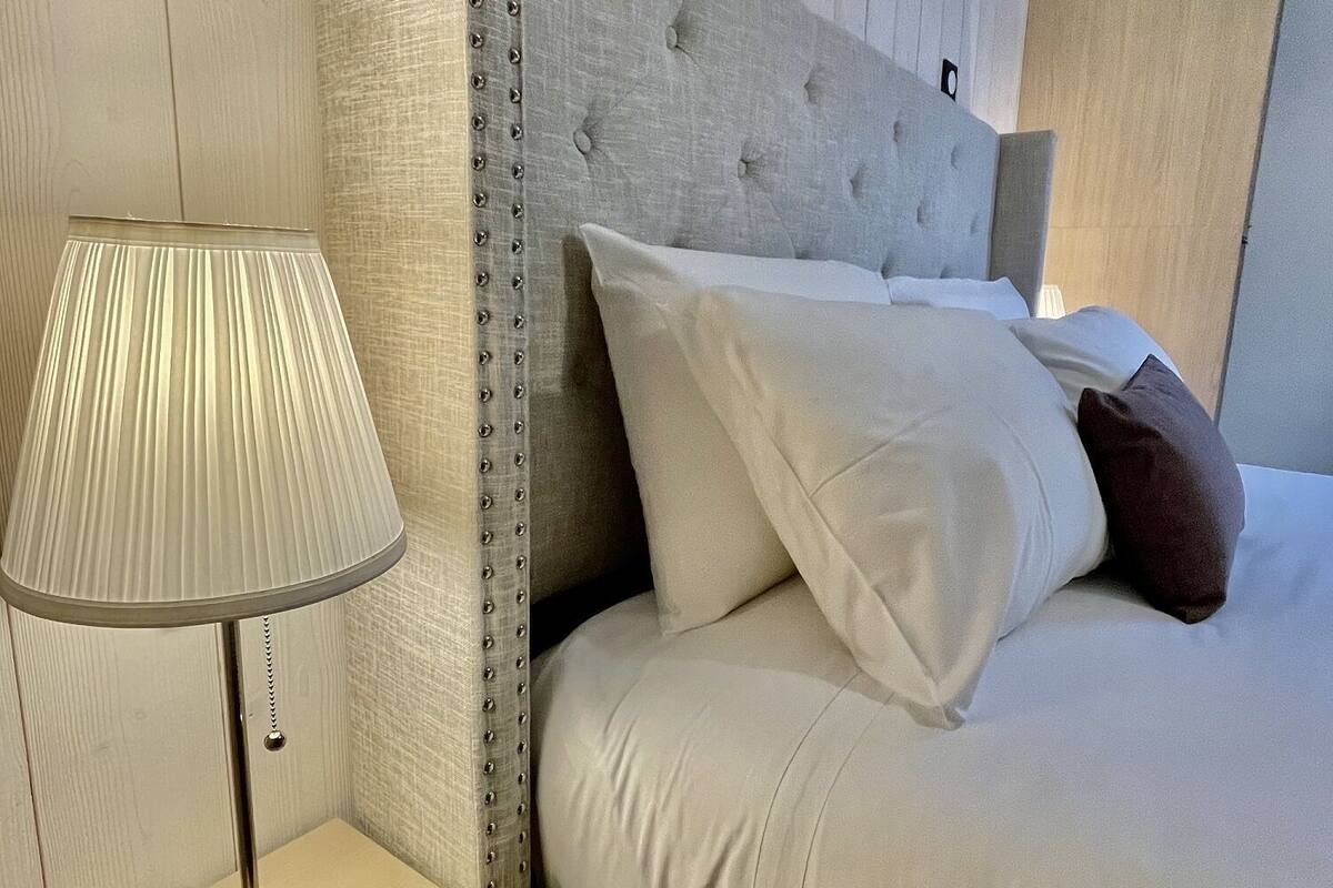 Cama bem arrumada com lençóis brancos, cabeceira com design acolchoado e luminária de cabeceira em mesa de madeira.