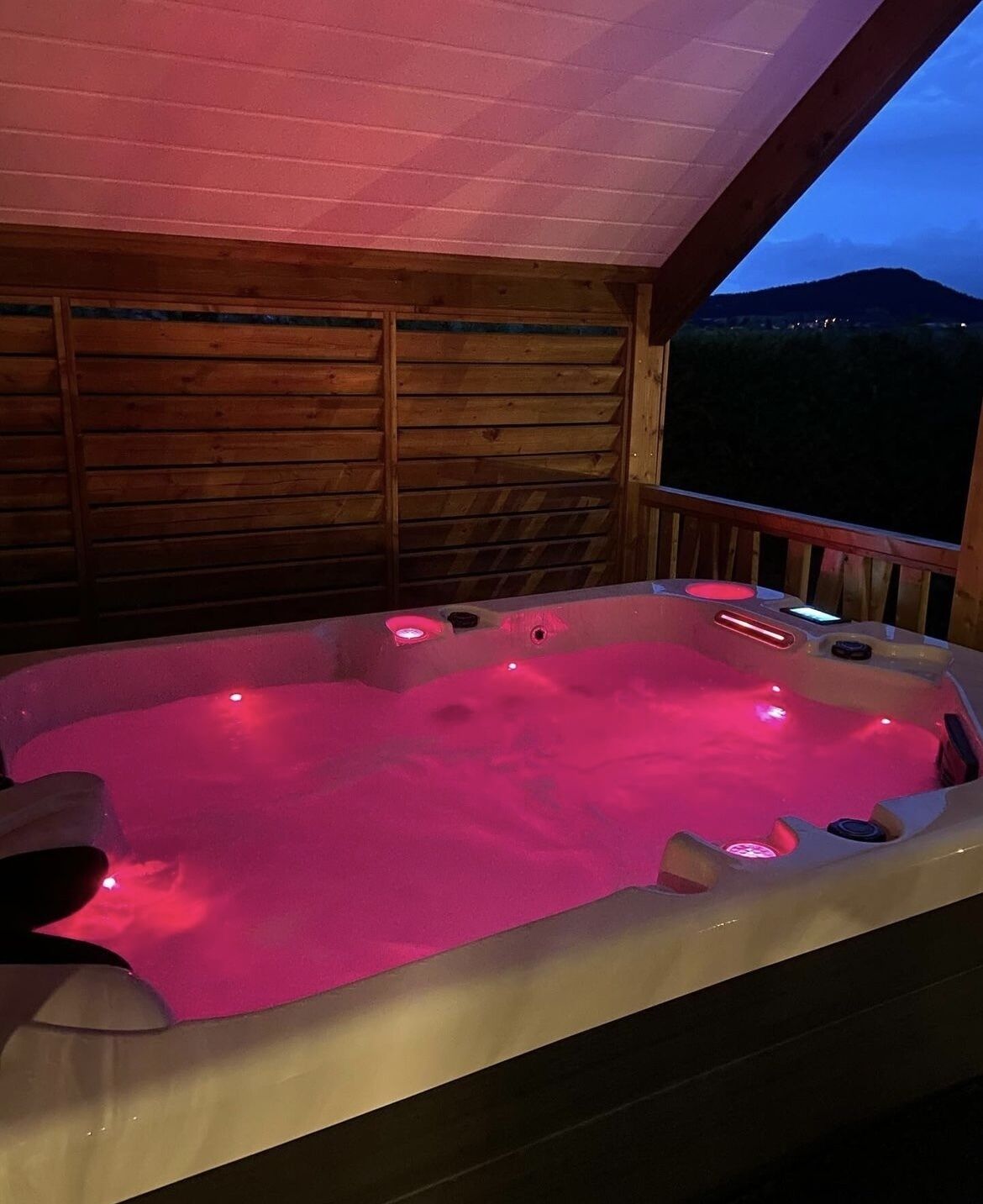Un jacuzzi con iluminación rosada, terraza de madera y vistas a montañas.