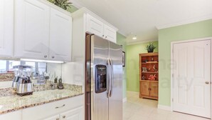 A kitchen with white cabinets, a stainless steel refrigerator, granite countertops, and a wooden cabinet with decorative items. - Dog Friendly Home with Ocean View from Balcony by RedAwning (Fernandina Beach)