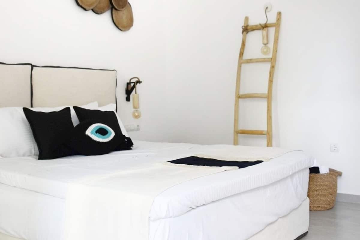 A minimalist bedroom with a large bed, white bedding, a wooden ladder, and a wall-mounted hat.