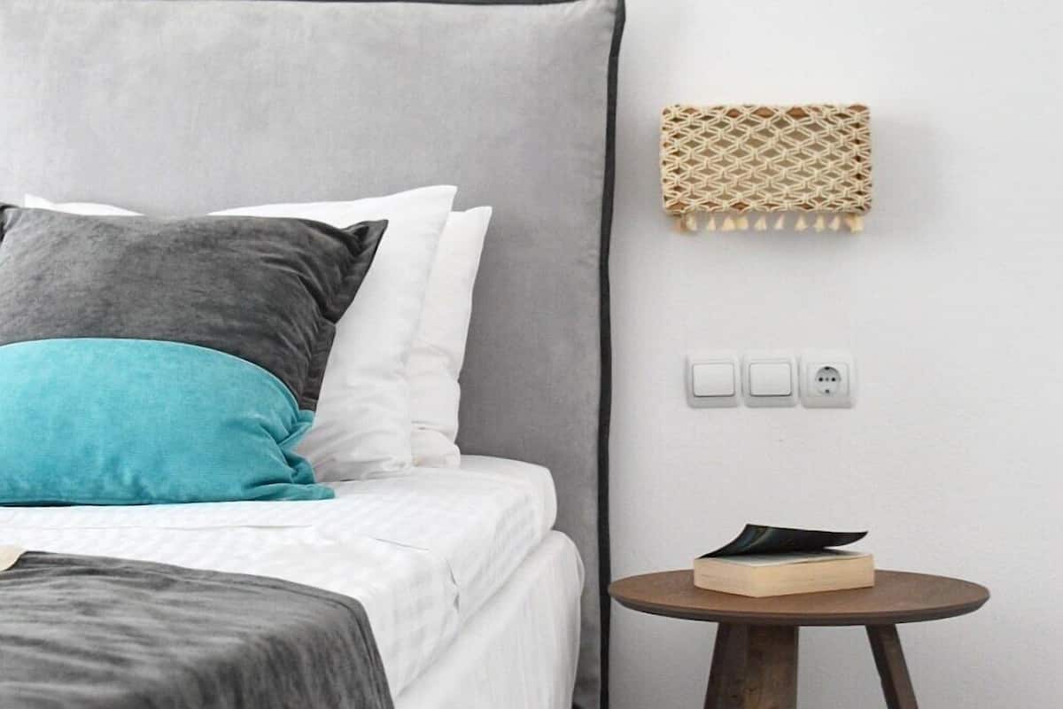 A bedroom with a grey upholstered headboard, a bed with white and teal pillows, a round wooden side table with a book, and a wall-mounted woven shelf.
