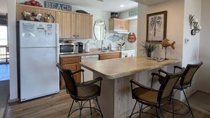 A compact kitchen with wooden cabinets, a refrigerator, a microwave, and a sink. There are two bar stools and a small dining table.