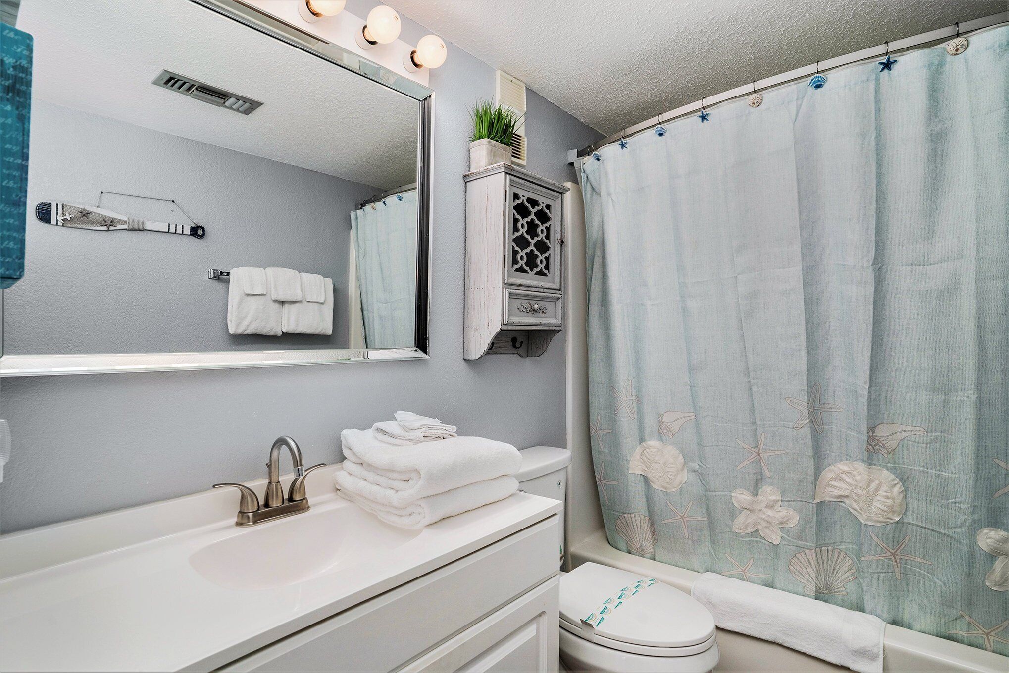 A bathroom with a white sink, a mirror with lights, a cabinet, and a shower curtain with a floral pattern.