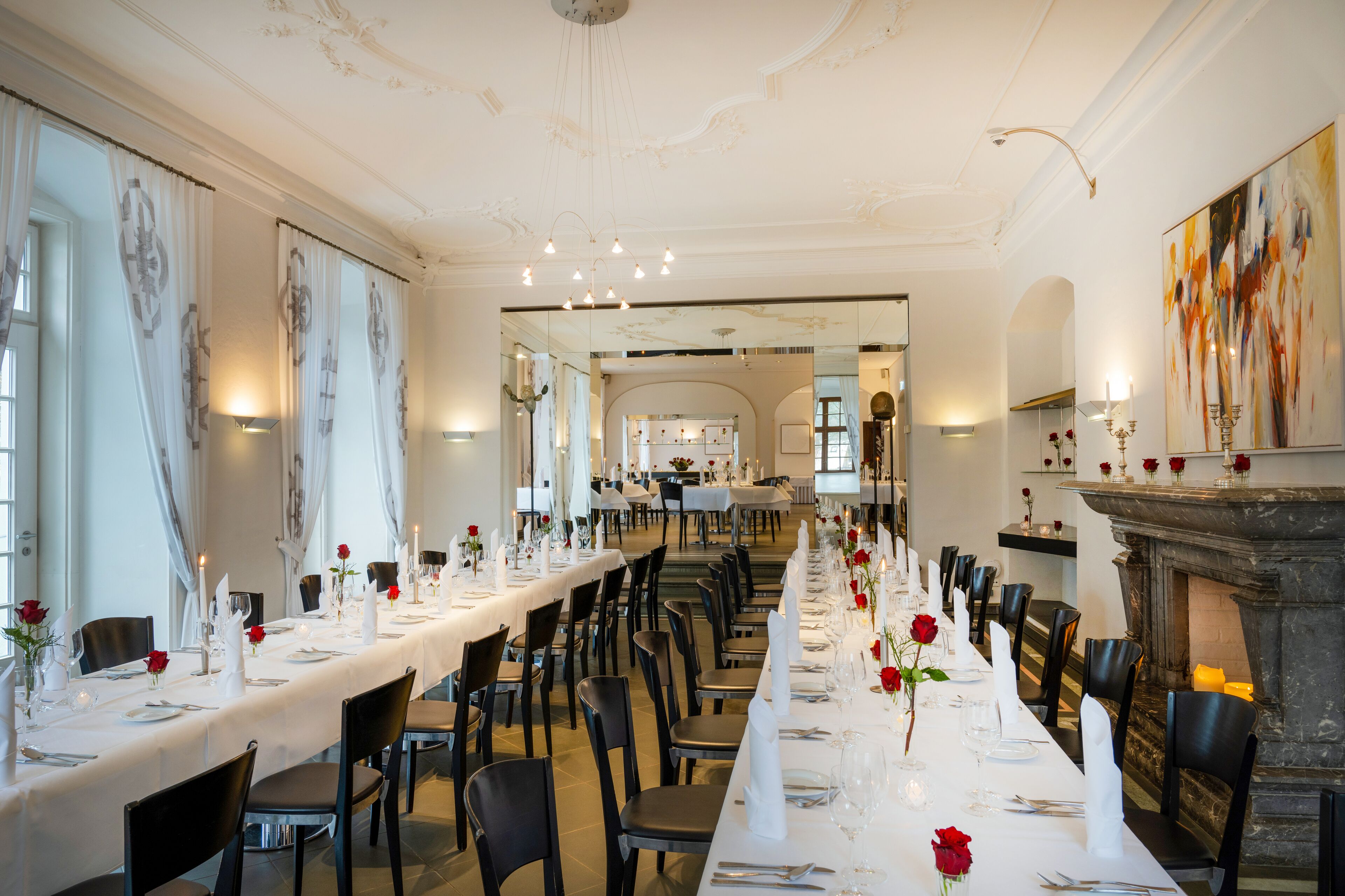 A long dining table set with white tablecloths, candles, and red roses. There are chairs arranged around the table, and a fireplace is visible in the background.