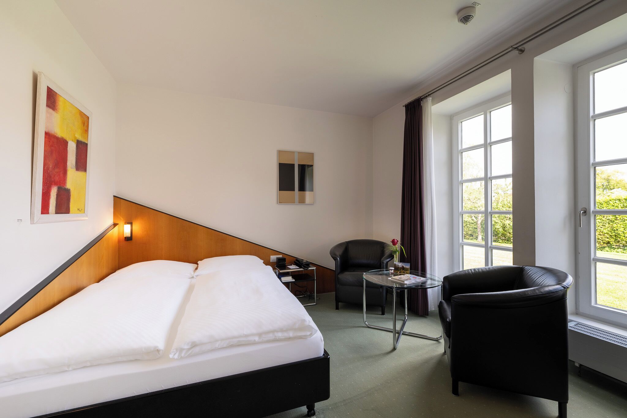 A hotel room with a bed, a desk, a chair, and a window with a view of greenery.