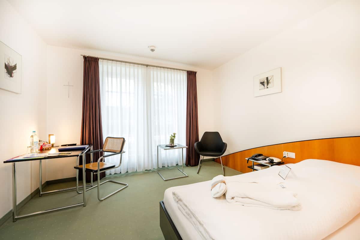 Een hotelkamer met een bed, een bureau, twee stoelen, een kleine tafel en een raam met gordijnen.